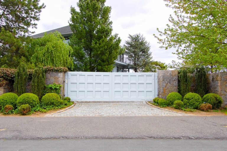 A blue gate protects a property with mature trees and lots of greenery. A blue house is slightly visible behind it.