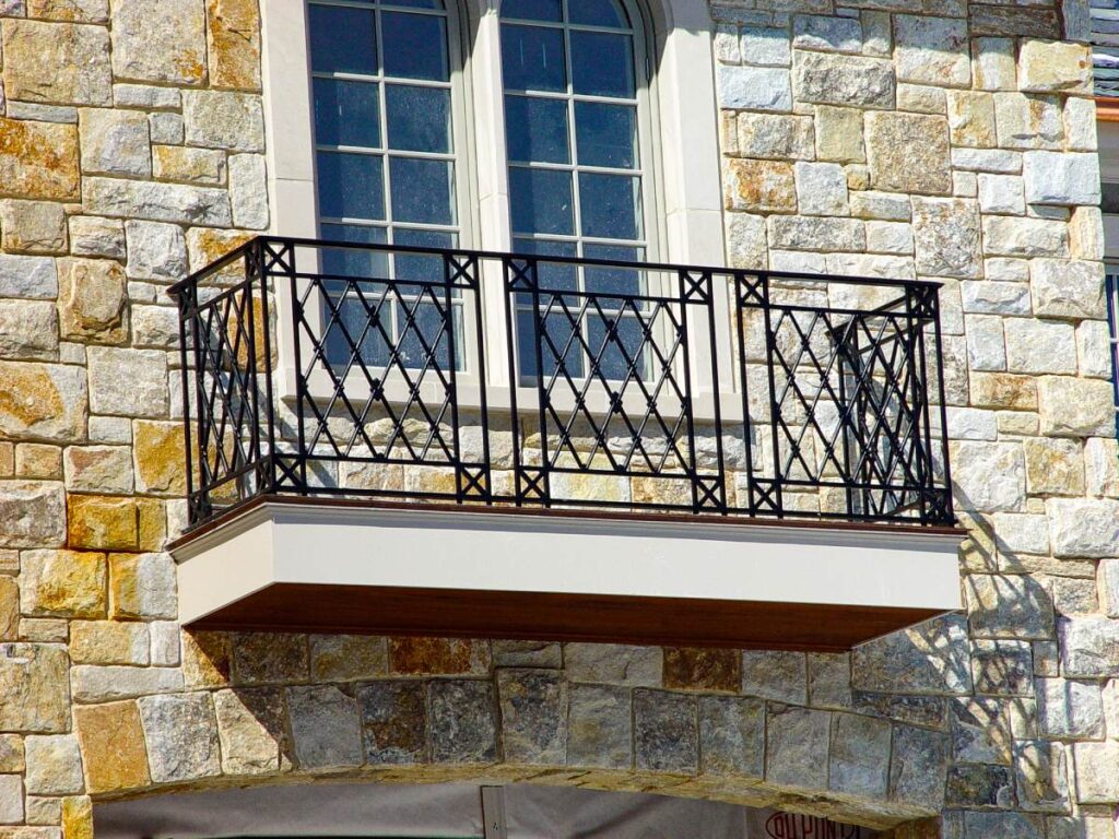 A black metal balcony with a criss-crossed geometric pattern outside a home’s stone exterior.
