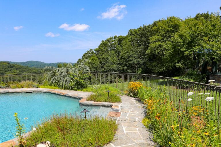 A bronze-colored wrought iron metal pool fence with ornate details on the pickets and below the railing encircles a pool overlooking a scenic view.