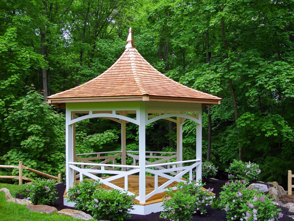 White garden gazebo with a cedar shingle roof surrounded by greenery