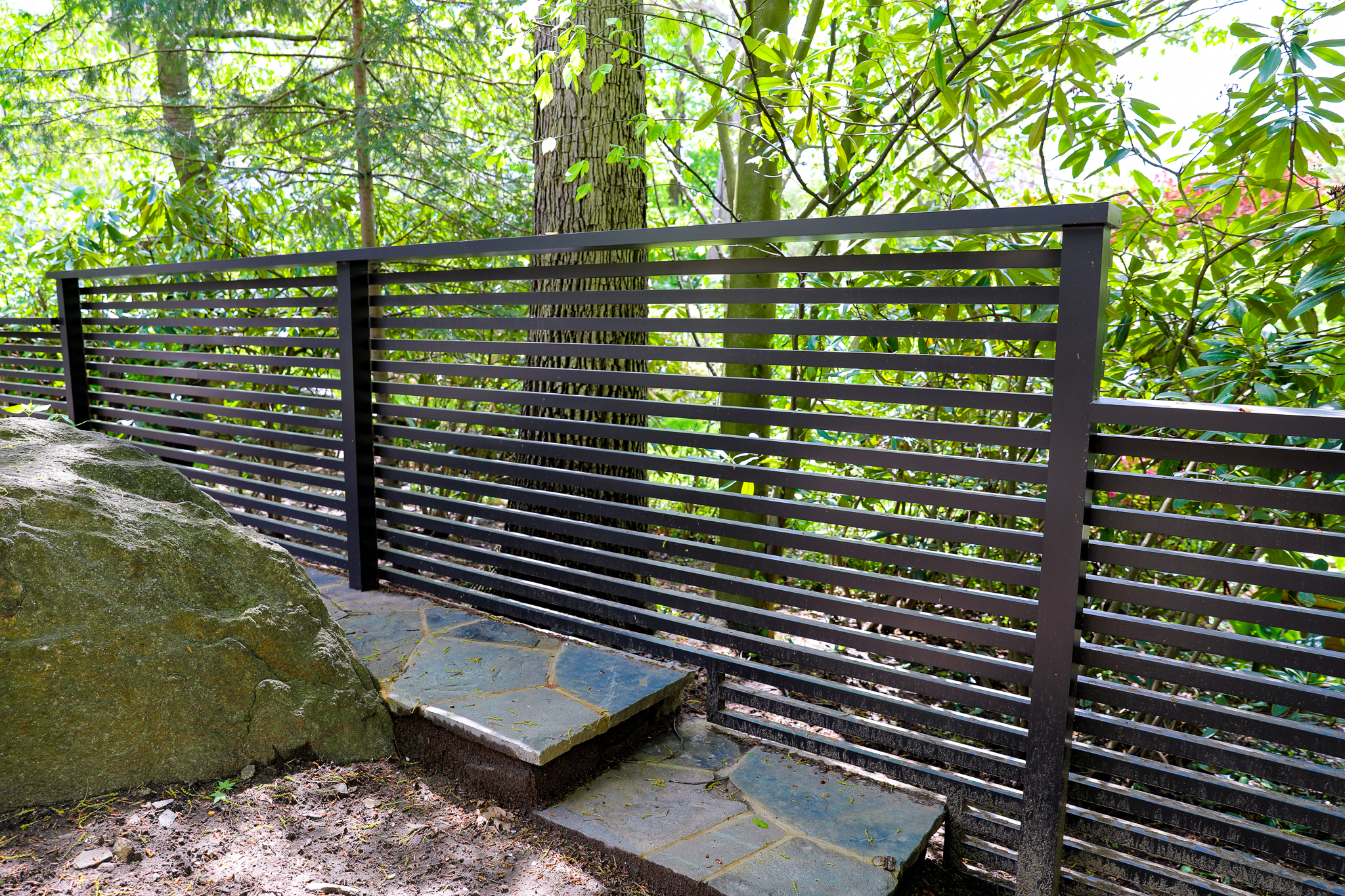 Modern black horizonal fence made of metal with a lush woods backdrop